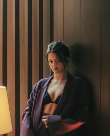 Attractive woman in oversized purple blazer and black bra poses indoors against a striped wood wall, with a table lamp visible.