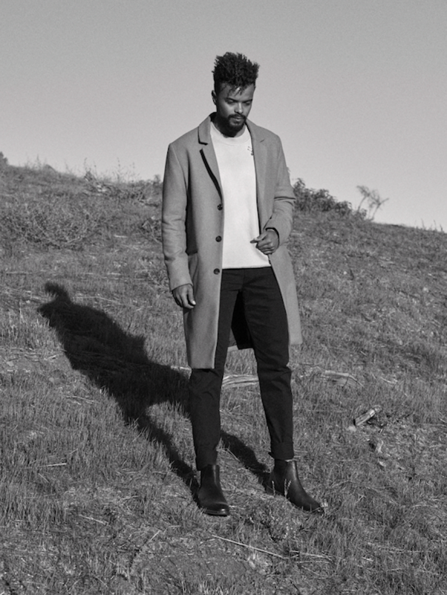 Black and white editorial portrait of a man standing on a grassy hill wearing a long coat and dark trousers.