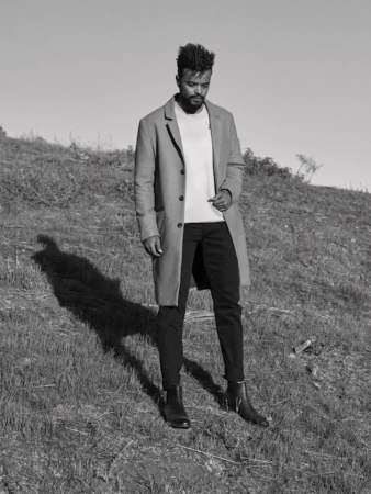 Black and white editorial portrait of a man standing on a grassy hill wearing a long coat and dark trousers.