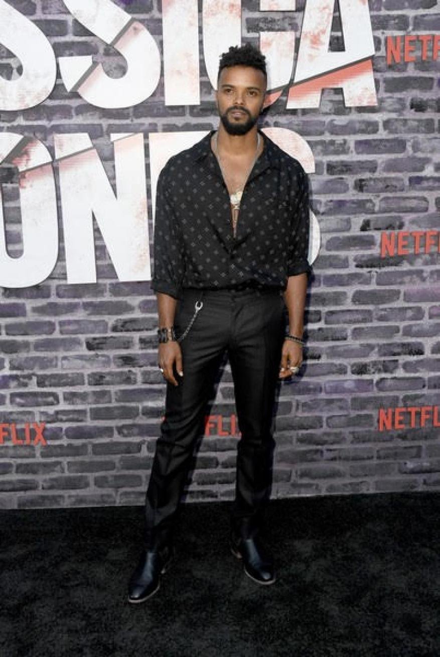 Full-length shot of a Black male in a patterned shirt and black pants on a black carpet, posing in front of a gray brick wall.