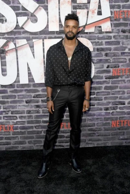 Full-length shot of a Black male in a patterned shirt and black pants on a black carpet, posing in front of a gray brick wall.