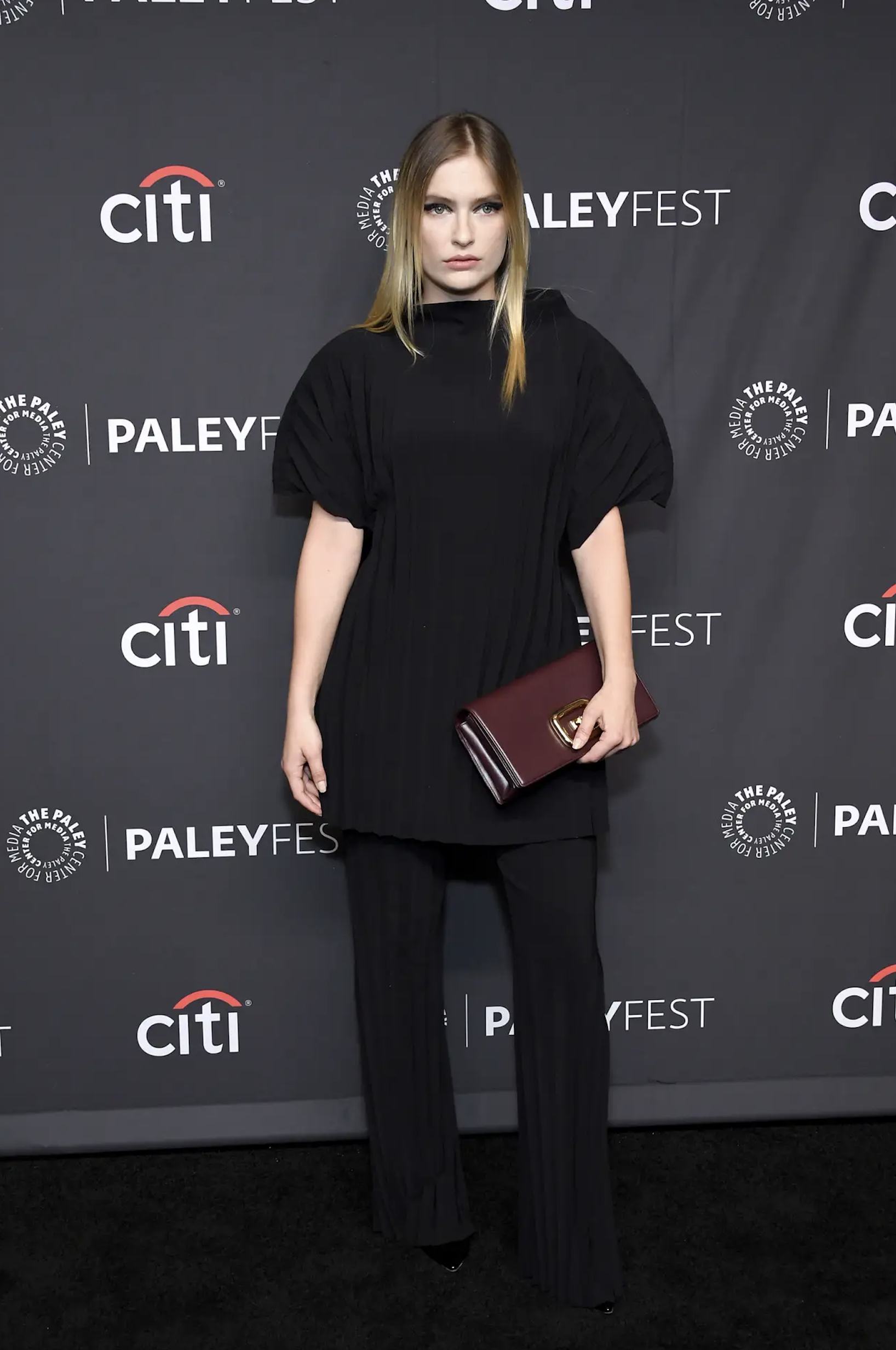 A woman in a black dress and pants poses with a maroon clutch at a PaleyFest event, with a black patterned backdrop and Citi logo.
