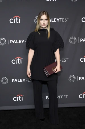 A woman in a black dress and pants poses with a maroon clutch at a PaleyFest event, with a black patterned backdrop and Citi logo.