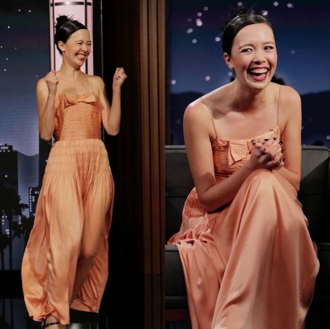 A woman with updo poses in a light orange dress on TV show set, smiling with hands clasped in front, captured in a medium shot.