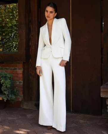 Elegant woman in a white suit posing against a wooden door backdrop, exuding sophistication and style in a bright, outdoor setting.