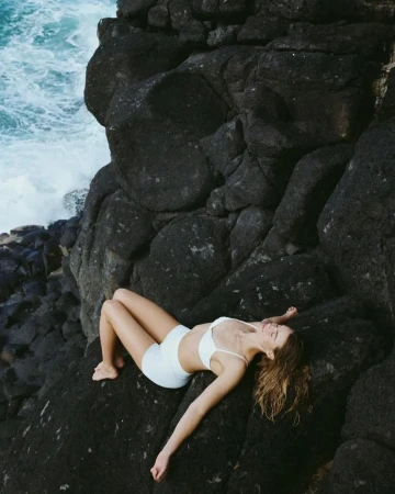 Woman wearing white swimwear lying on dark volcanic rock beside crashing ocean waves
