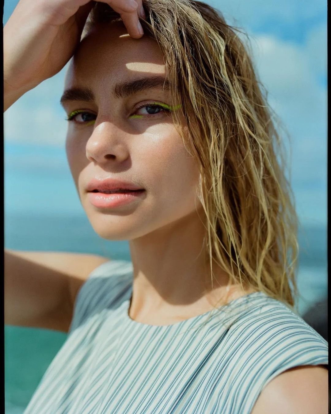 Close-up portrait of a blonde woman with wet hair and bright yellow eyeliner standing outside with a blue ocean background on a sunny day.