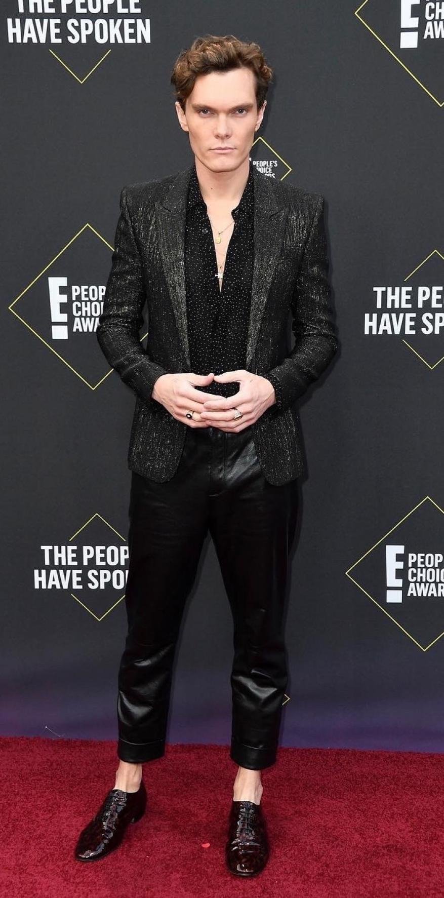 Full-length shot of a Caucasian adult male in a dark blazer, pants, and shoes, posing on a red carpet with branding in the background.