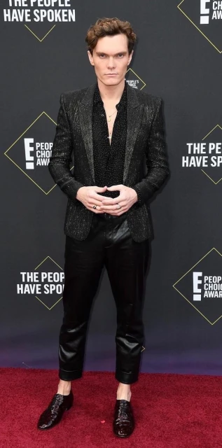 Full-length shot of a Caucasian adult male in a dark blazer, pants, and shoes, posing on a red carpet with branding in the background.