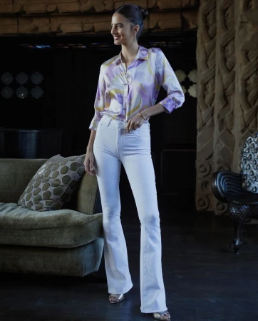 A woman with her hair up poses in a buttoned swirl-patterned blouse and white flared pants in a dimly lit indoor setting.