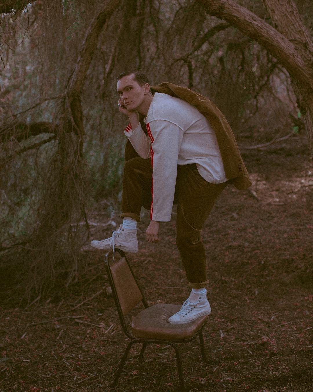 Young man balancing on a chair outdoors wearing a white long-sleeve shirt, brown pants, a brown jacket, and white sneakers in a forest setting.