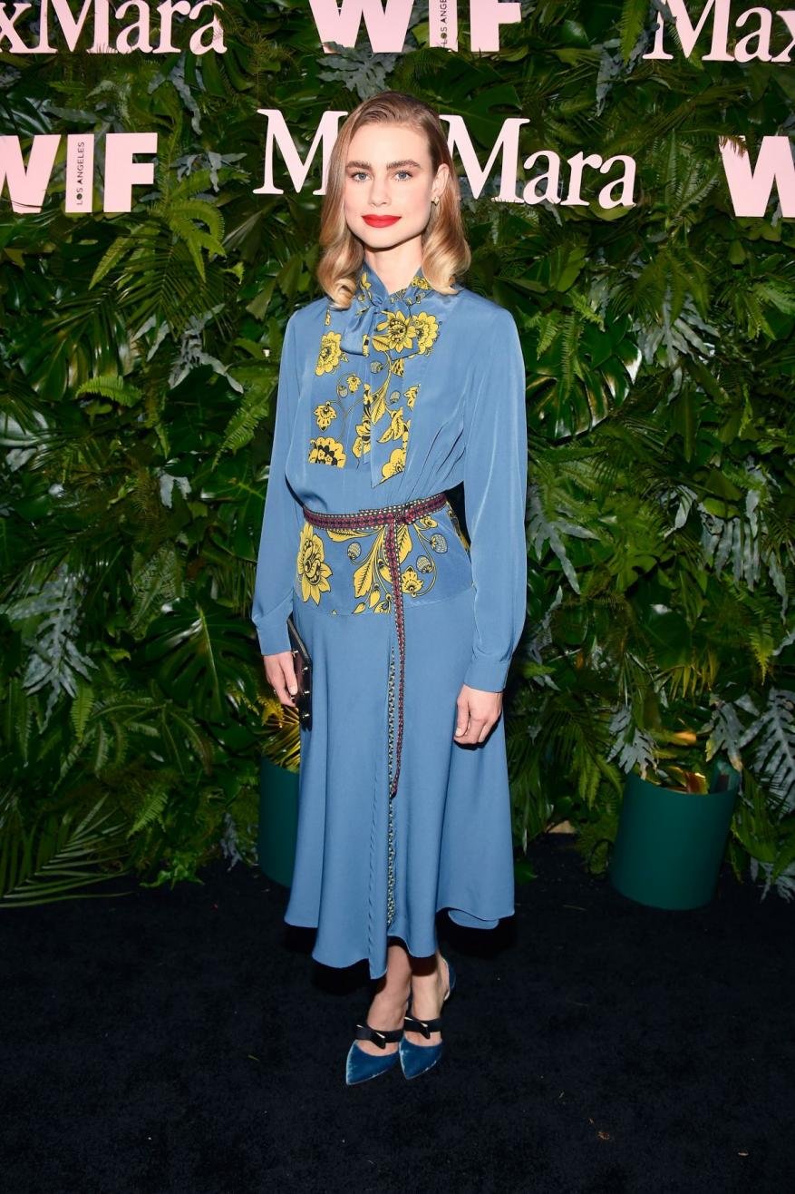 Woman poses in a blue dress with floral accents against a backdrop of green foliage at an event, showcasing fashion and style.