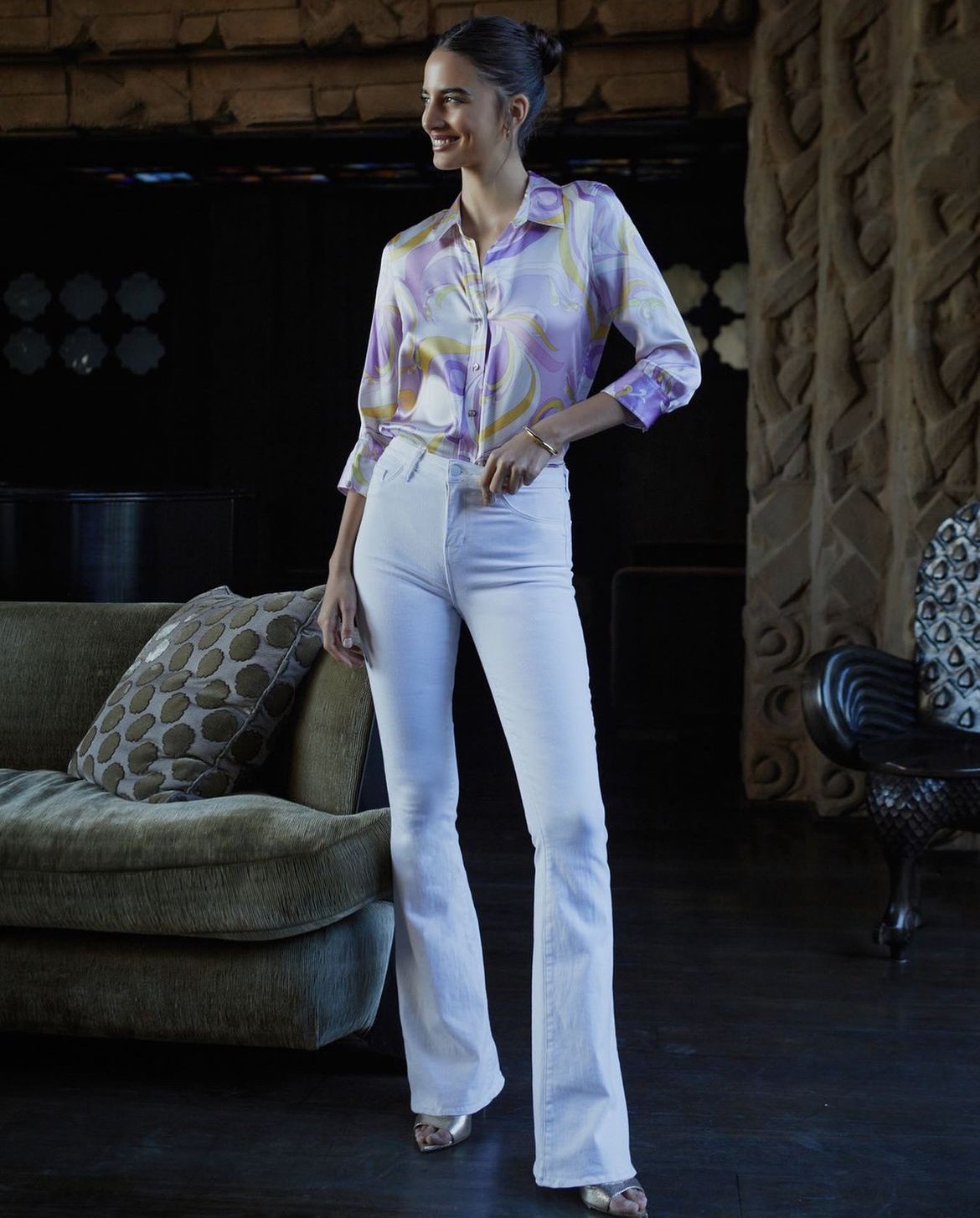 A stylish woman in a patterned blouse and white jeans poses in an indoor setting, with a dark background and a textured sofa.