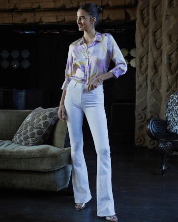 A stylish woman in a patterned blouse and white jeans poses in an indoor setting, with a dark background and a textured sofa.