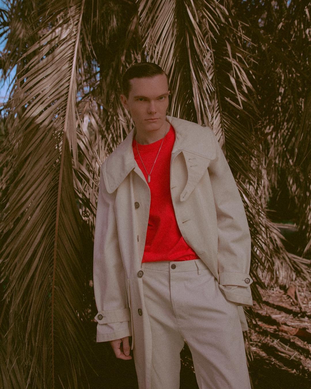 Man in neutral trench coat and red shirt posing near palm tree in bright outdoor light looking at camera in fashion editorial.