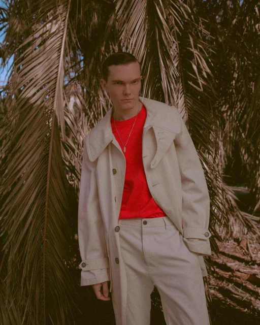 Man in neutral trench coat and red shirt posing near palm tree in bright outdoor light looking at camera in fashion editorial.