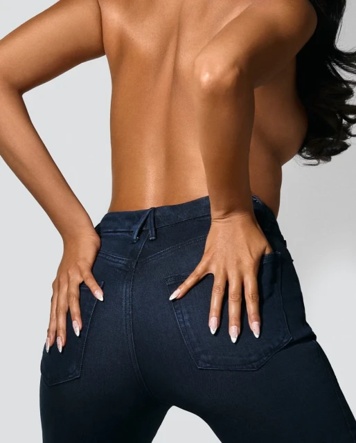 A woman poses from the back, hands on her hips wearing tight, dark wash jeans against a bright background in a studio fashion shot.