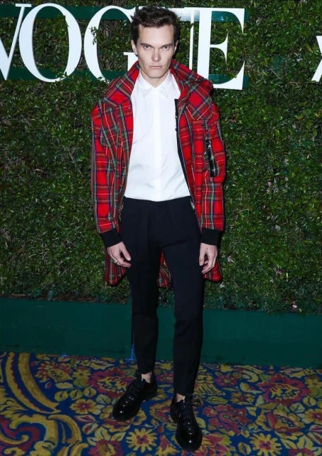 Adult male in plaid coat and black pants stands in front of green leafy wall.  The word 'Vogue' is visible in the upper left.