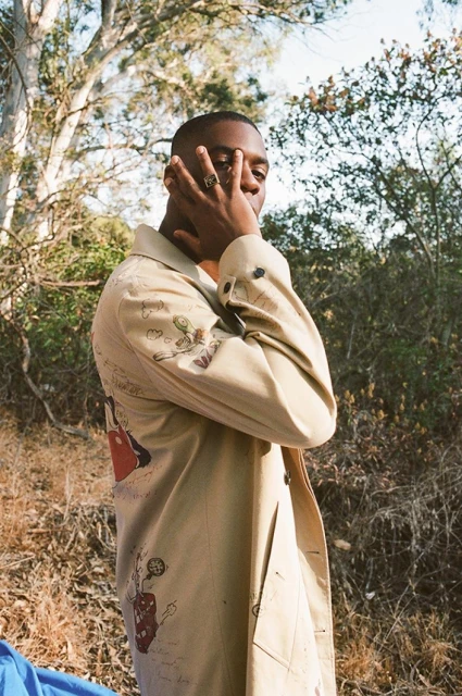 Young man in patterned beige trench coat stands outdoors with trees. His hand is raised to partially cover his face with jewelry.