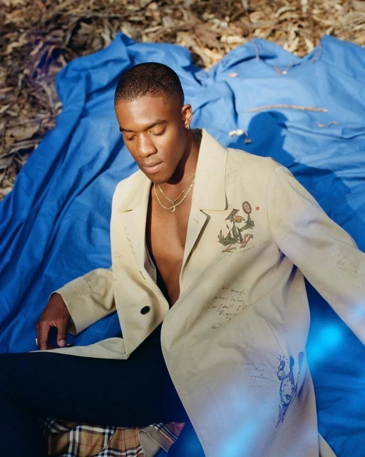 Portrait of adult male model wearing a tan trench coat with drawings, sitting on blue and plaid fabric outdoors in natural light.