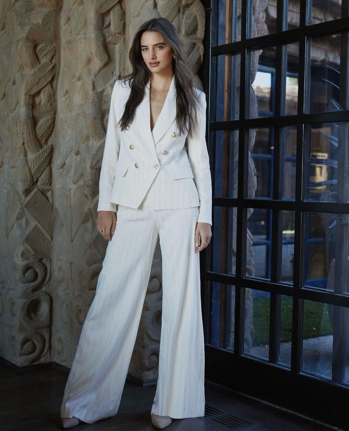 Elegant woman in a white double-breasted suit, posing against an ornate wall and dark framed window in a fashion forward style.