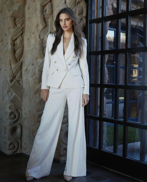 Elegant woman in a white double-breasted suit, posing against an ornate wall and dark framed window in a fashion forward style.