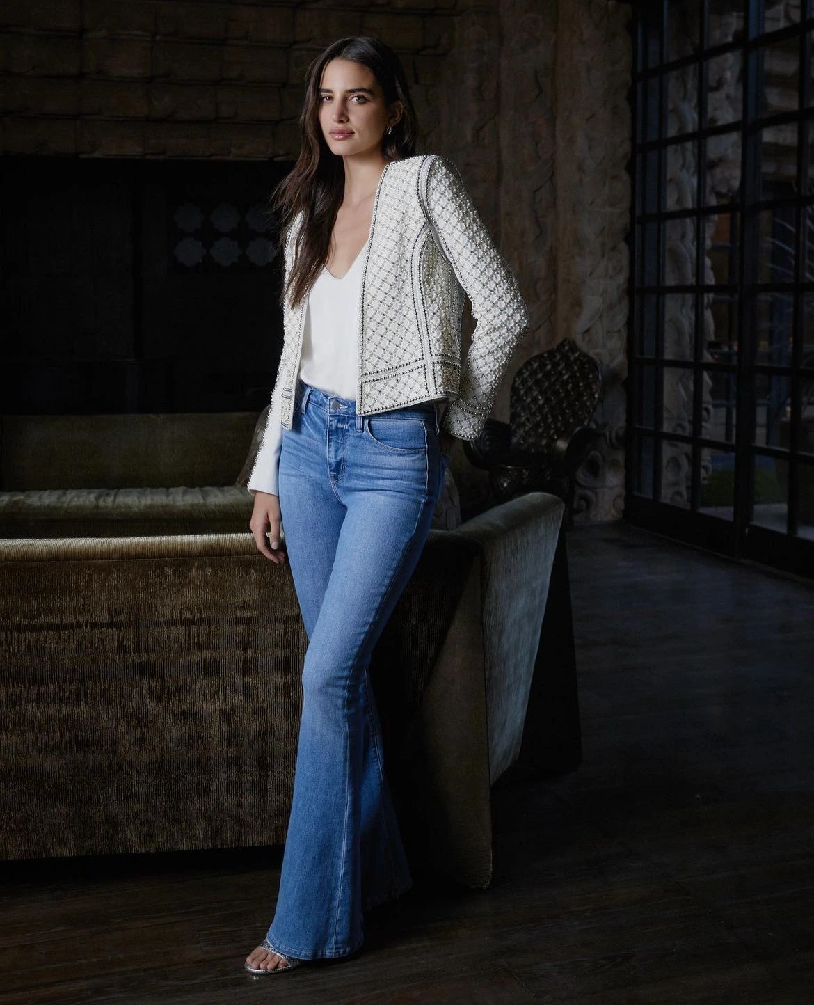 Woman in jeans and a jacket poses indoors, near a couch. The shot captures fashion and lifestyle in a staged setting.