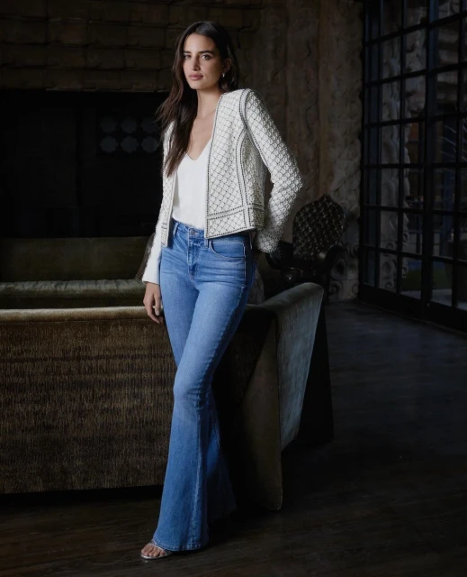 Woman in jeans and a jacket poses indoors, near a couch. The shot captures fashion and lifestyle in a staged setting.