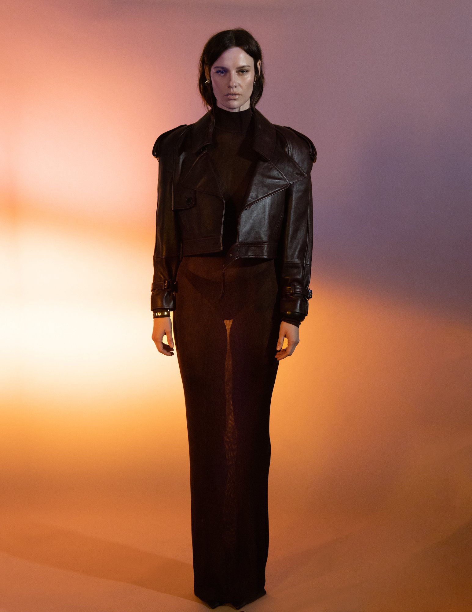 A full-length studio portrait of a woman with dark hair wearing a black leather jacket and long black skirt against an orange-purple gradient background.