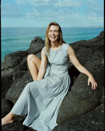 Smiling woman in a striped sleeveless dress seated on dark volcanic rocks near the ocean.