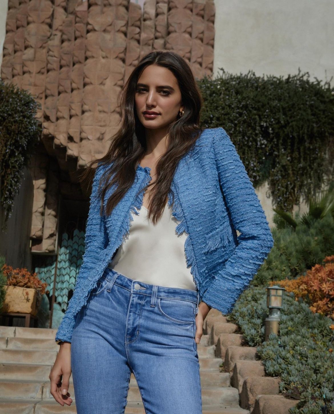 A brunette woman models a blue tweed jacket with a cream-colored top and blue jeans against an outdoor background with stone steps.