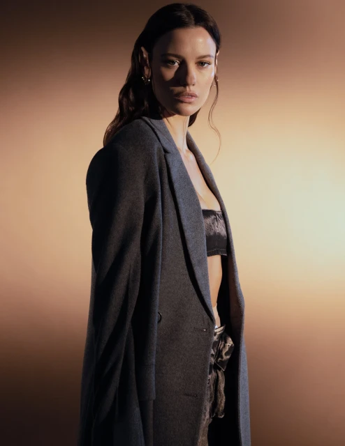 Fashion portrait of a woman in a grey coat and bra top, posing in a photo studio with warm-toned lighting.