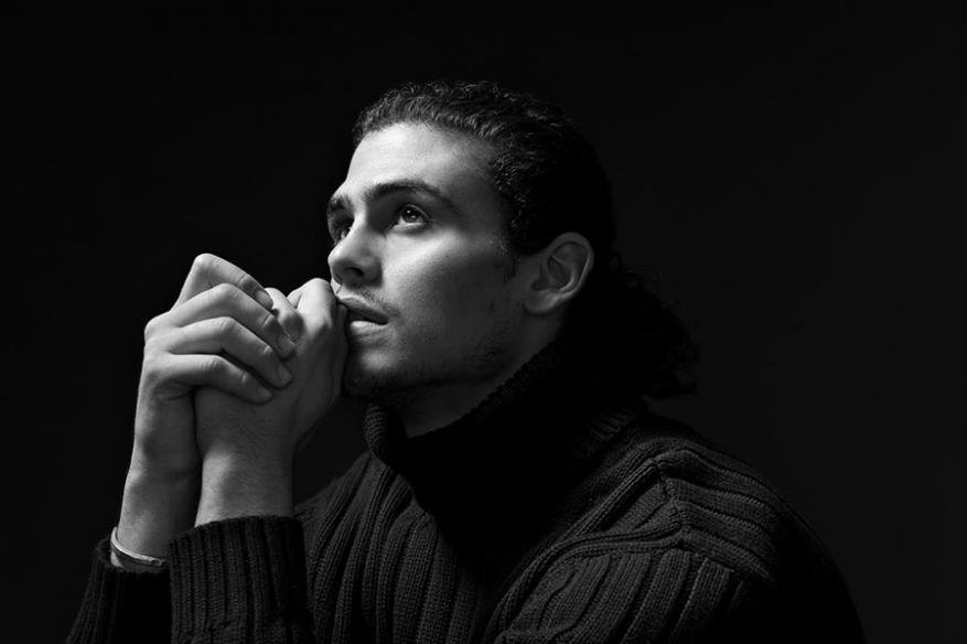 Black and white portrait of a young man in a black turtleneck sweater, looking upwards with his hands clasped under his chin.