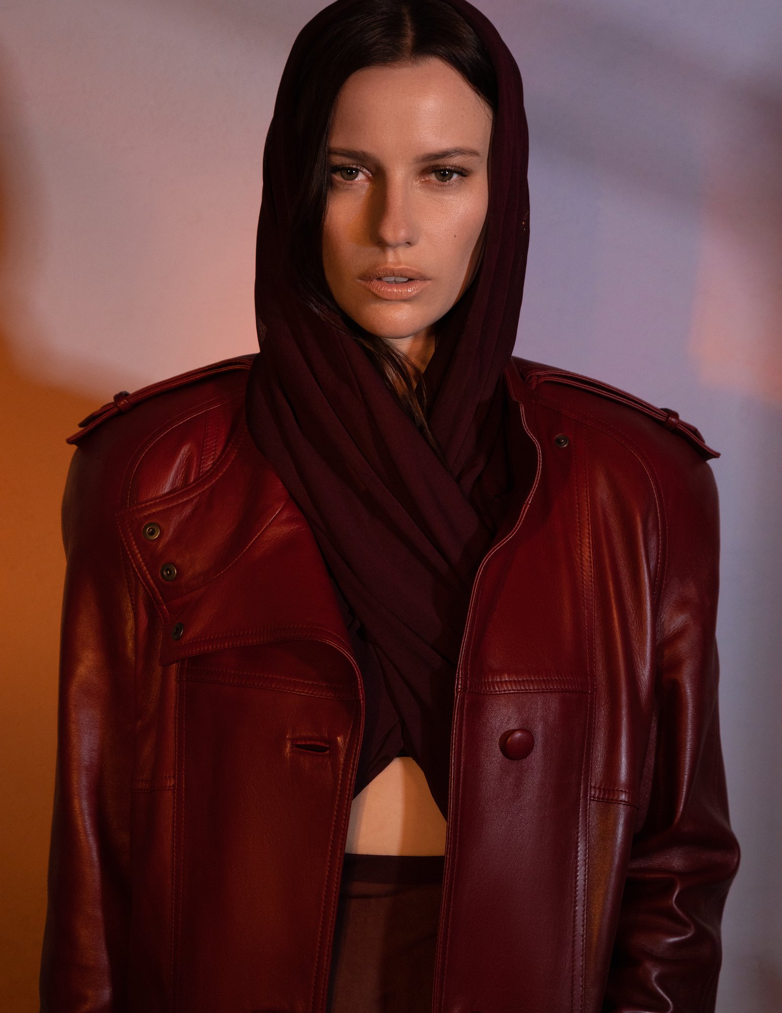 Portrait of an adult woman wearing a burgundy leather jacket and hooded top, posing in a photo studio with soft lighting.