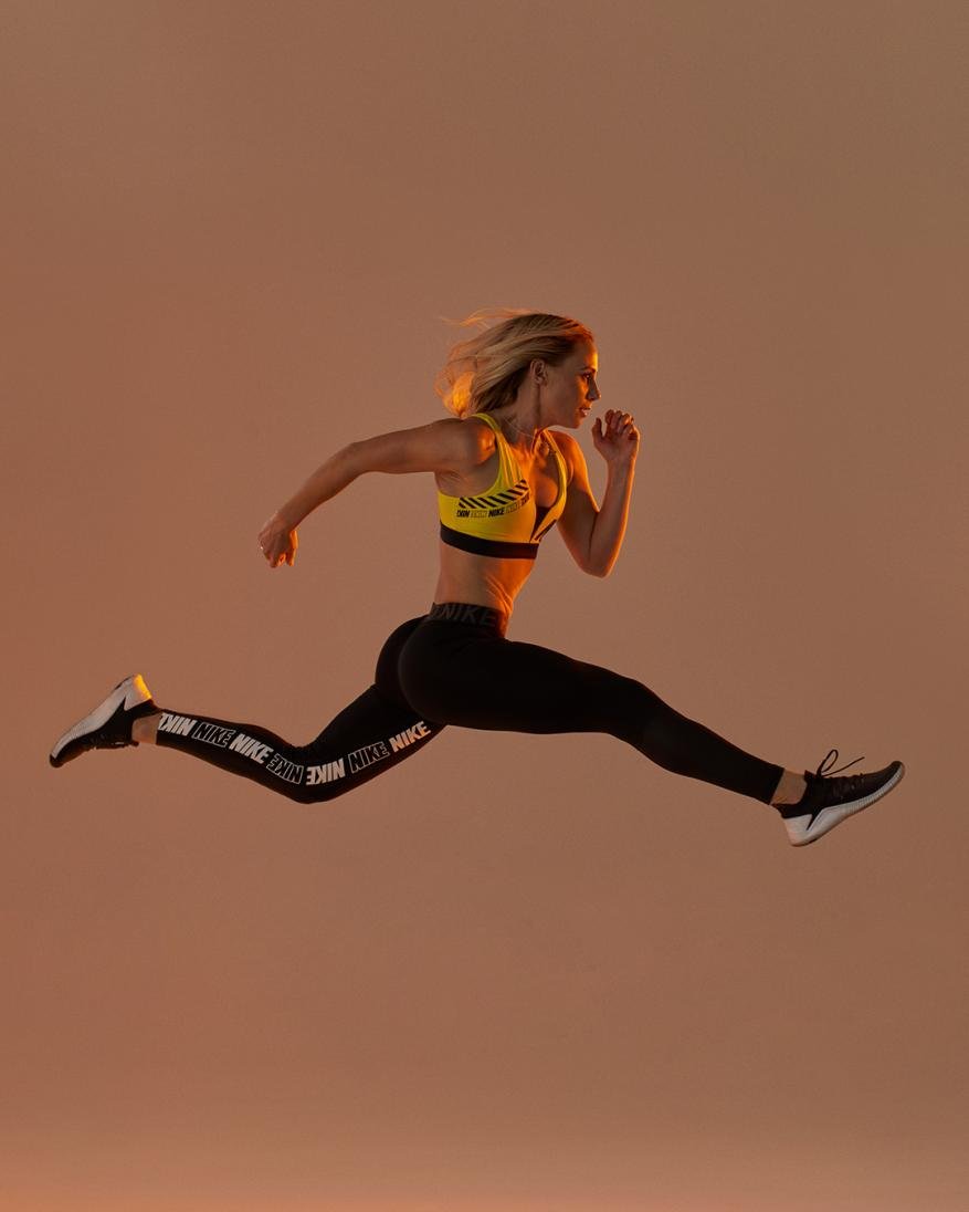 Blonde woman in yellow sports bra and black Nike leggings leaps in a mid-air running pose against a warm-toned peach background.
