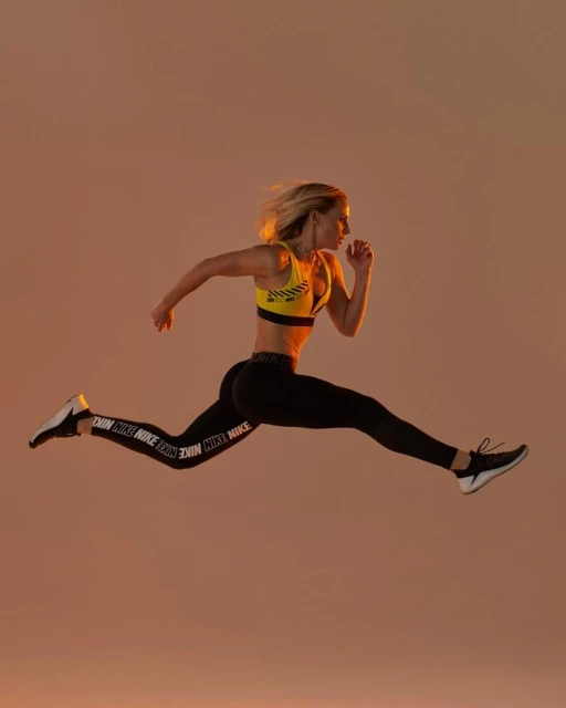 Blonde woman in yellow sports bra and black Nike leggings leaps in a mid-air running pose against a warm-toned peach background.