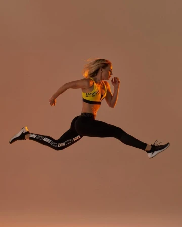 Blonde woman in yellow sports bra and black Nike leggings leaps in a mid-air running pose against a warm-toned peach background.