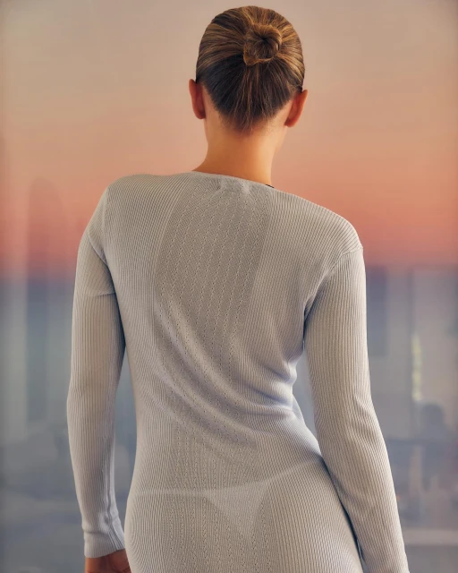 Back view of a fair-skinned adult woman with a high bun, wearing a form-fitting, long-sleeved dress against a blurred background. 