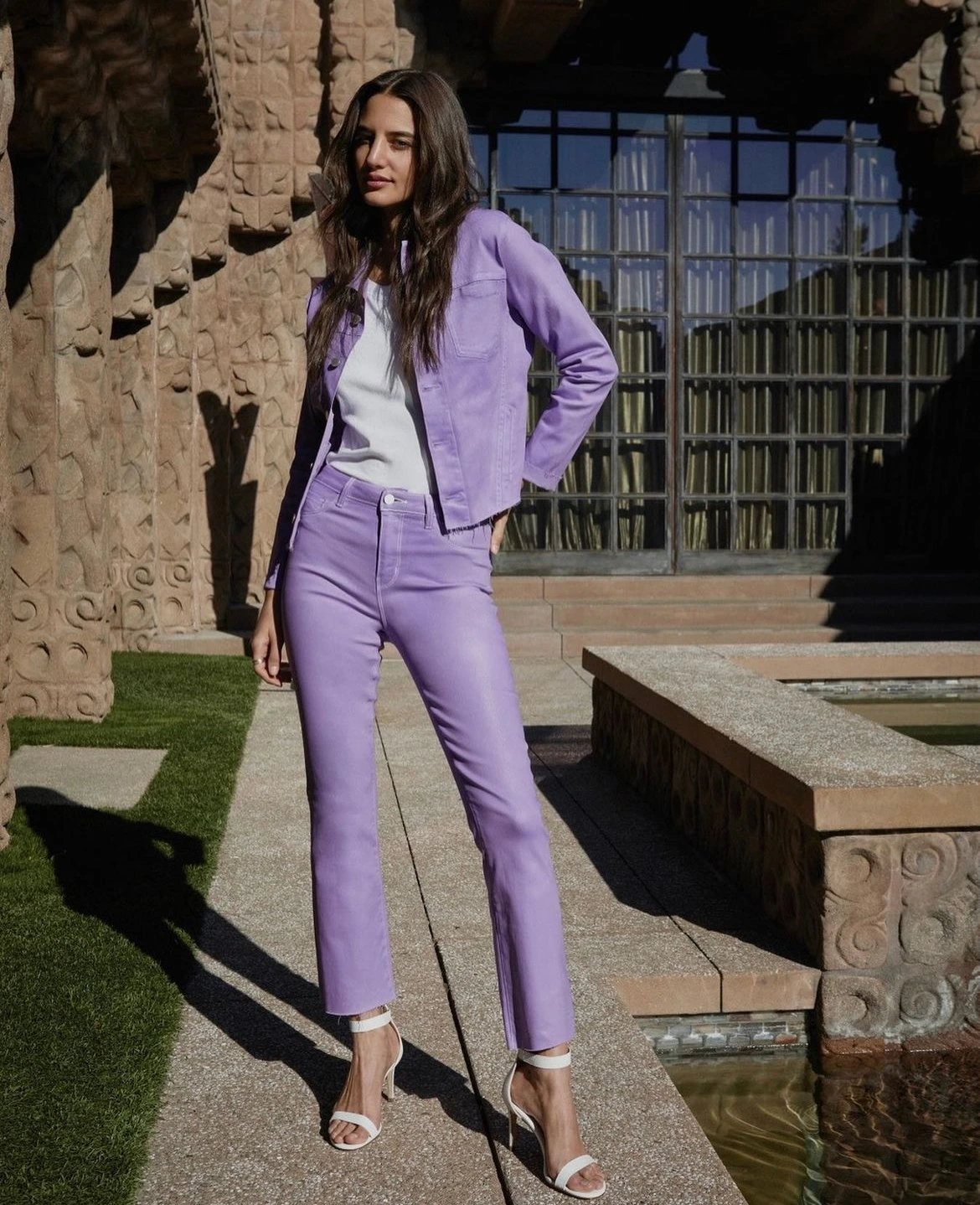 A long-haired woman poses outside in a matching purple jacket and flared pants, accessorized with white heels near a stone building.