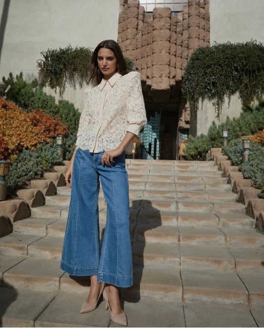 A woman poses on stone stairs in wide-leg jeans and a lace blouse, set against an architecturally detailed building and surrounding greenery.