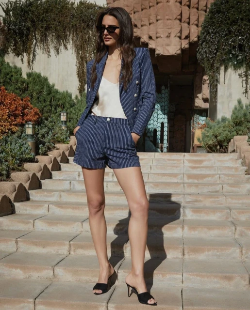A brunette woman models a pinstripe blazer and shorts set on stone steps, accessorized with black sunglasses and heeled mules, outdoors.