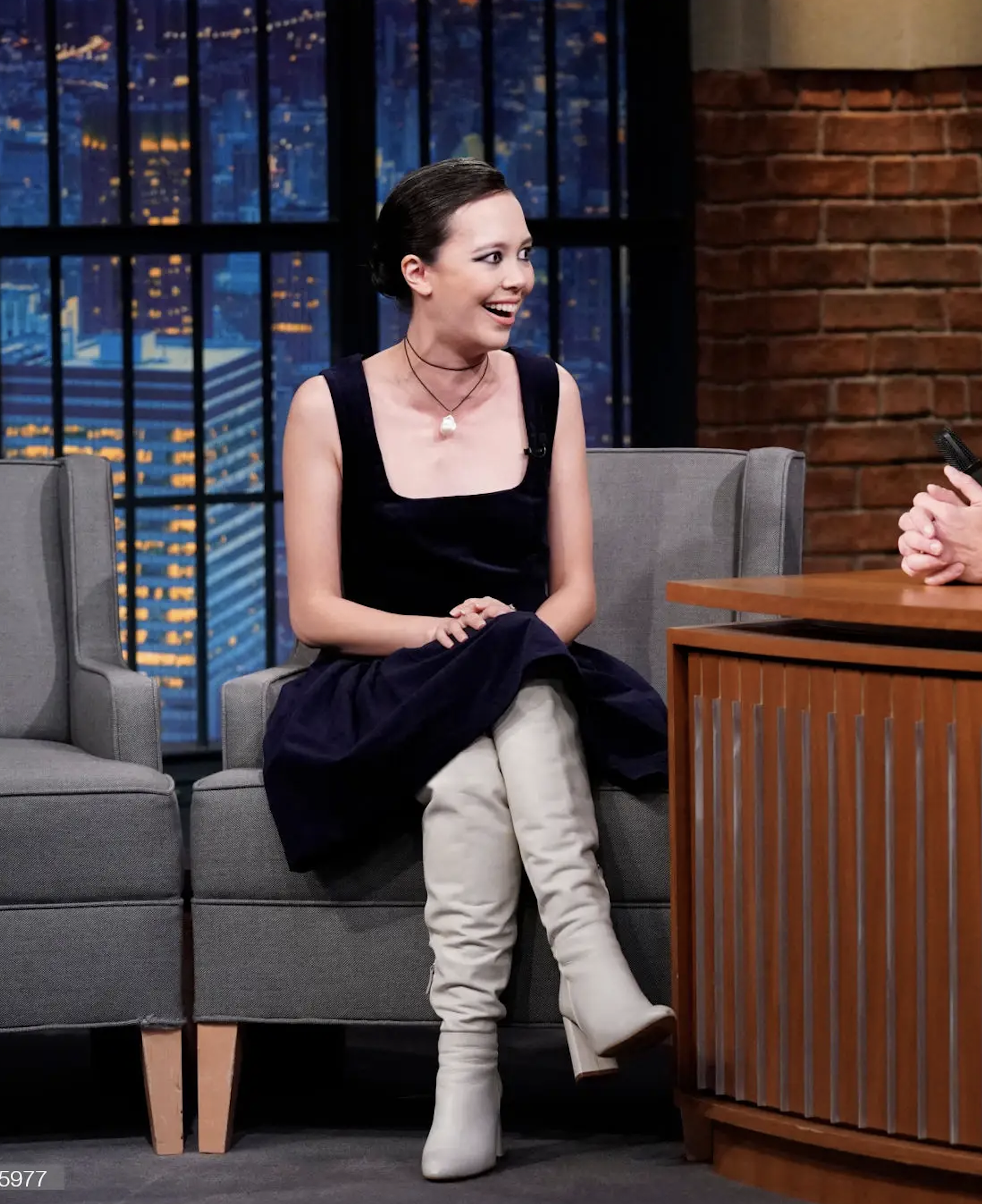A woman sits smiling on set with a dark dress and light-colored boots in a medium shot during what appears to be a late-night show.