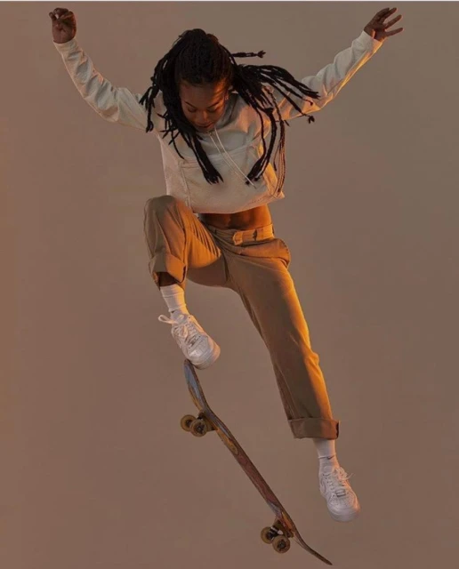 Young woman with dreadlocks in mid-air skateboarding; she wears a hoodie, chinos, and white sneakers against a neutral backdrop.