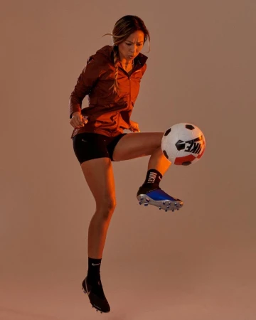 Young woman in sportswear dynamically juggling a soccer ball with her knee in a studio setting with soft, diffused lighting.