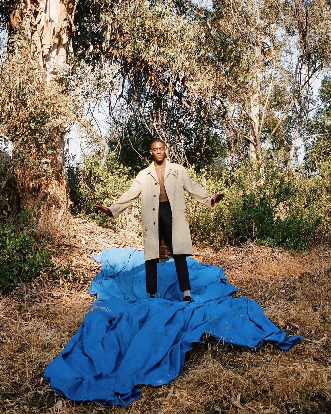 An adult Black man stands barefoot on blue fabric in a forest clearing, wearing black pants and an open tan trench coat, arms outstretched.