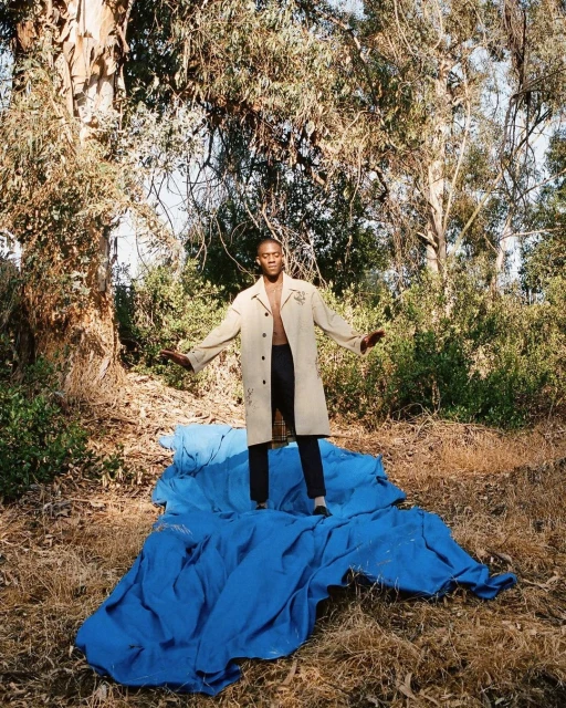 An adult Black man stands barefoot on blue fabric in a forest clearing, wearing black pants and an open tan trench coat, arms outstretched.