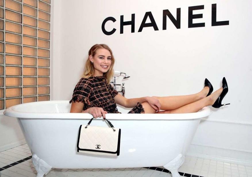 Blonde woman in a bathtub holding a Chanel purse, wearing black heels, and a tweed dress in a bright indoor setting.