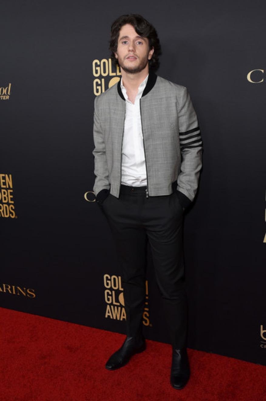 A man in a jacket and black pants poses on a red carpet in front of a wall with logos at an event with soft, diffused lighting.