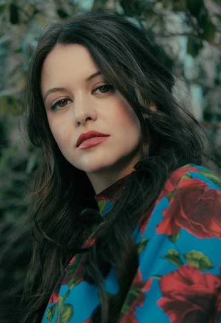 Portrait of a young adult female with long brown hair and a floral shirt, gazing directly at the camera against an organic green background.