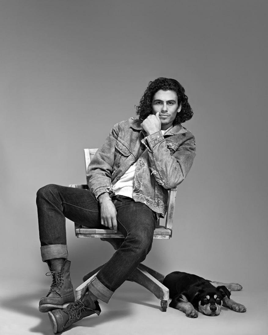 Monochrome portrait of a man with curly hair, wearing a denim jacket, seated on a chair next to a resting Rottweiler puppy.